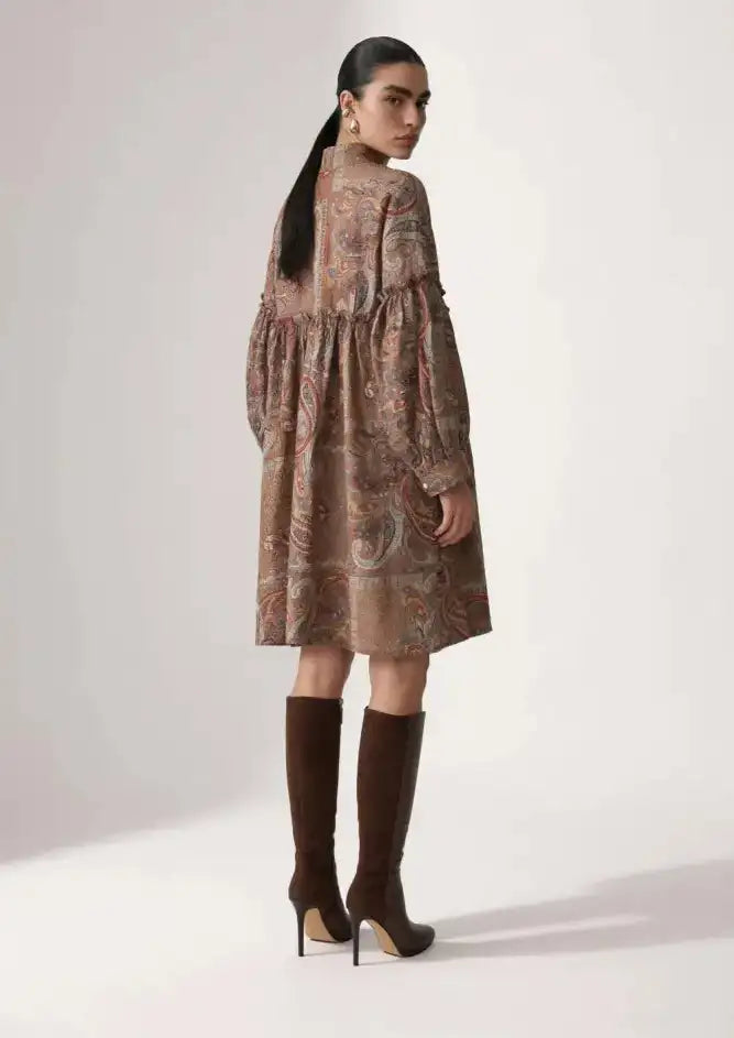 A woman wears a long-sleeved, knee-length dress with an intricate paisley pattern in earthy tones of brown and beige.